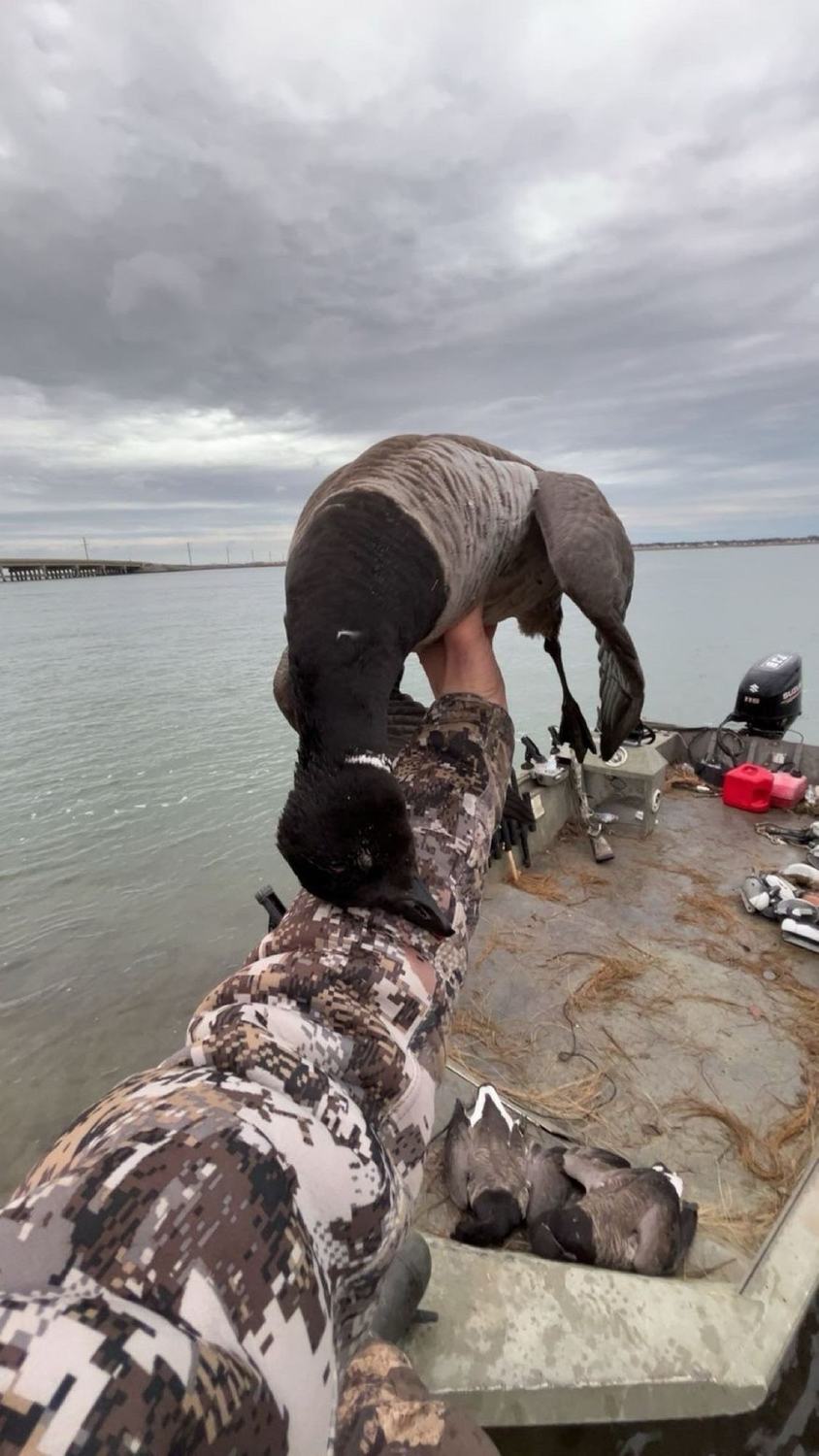 A hunter on the boat deck holding up a goose with open water and overcast sky behind