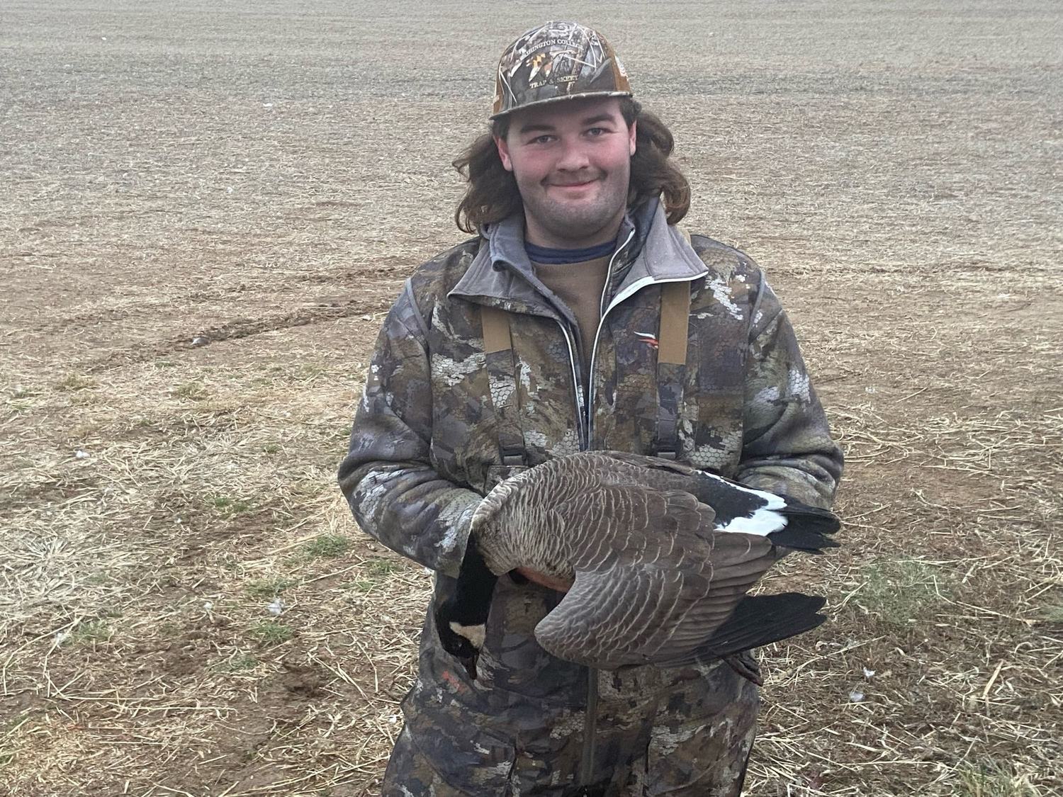 A hunter in camo holding a Canada goose in a cut field
